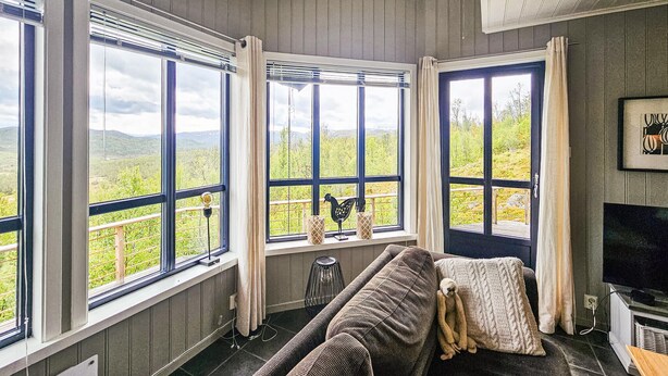 A room with large windows offering a view of a green landscape, a sofa, a television, and a decorative rooster figure. - Amazing home in Edland with sauna (Edland)