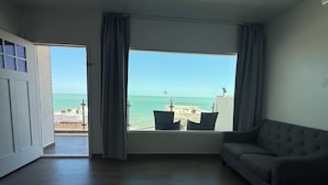 Een kamer met een groot raam dat uitzicht biedt op zee, een bank en een deur naar een buitenruimte.