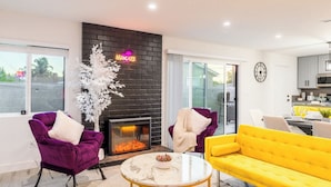 Ein modernes Wohnzimmer mit Kamin, violetten und gelben Sofas sowie einem durch eine Glastür einsehbarer Küchenbereich.