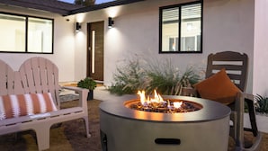 Ein Patio mit einer runden Feuerstelle, einem weißen Adirondack-Stuhl und einem Holzstuhl mit orangefarbener Sitzkissen.