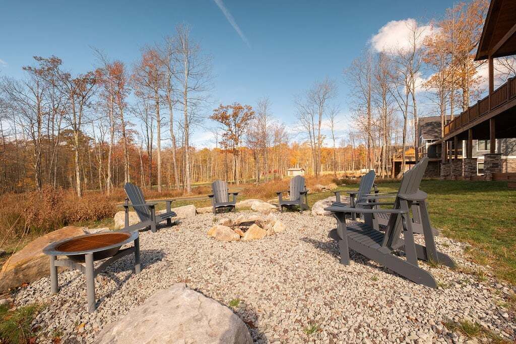 Een rustieke buitenruimte met Adirondack-stoelen en een vuurkuil, omgeven door grind en bomen.