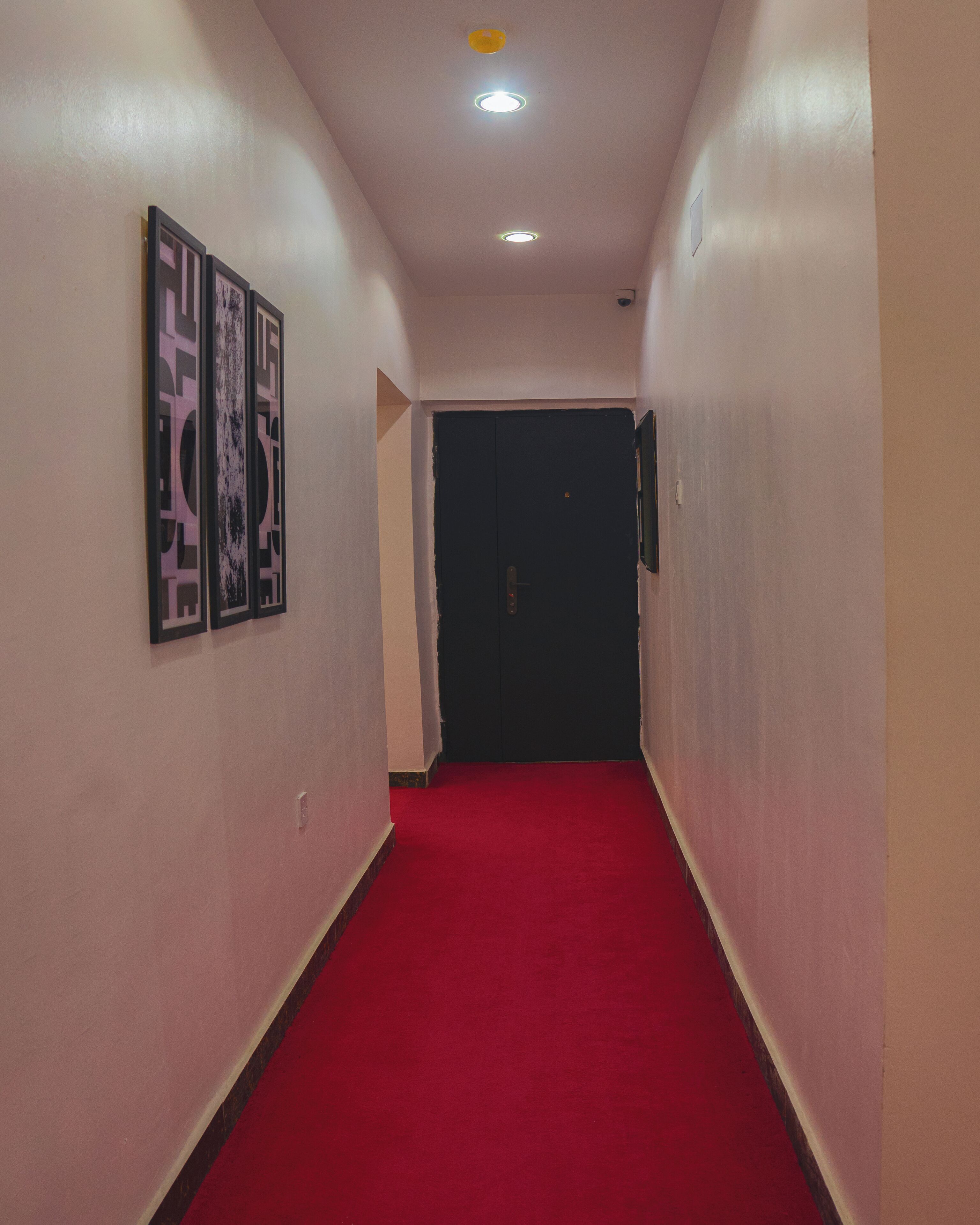 Un pasillo con alfombra roja, una puerta negra al final y tres cuadros en la pared.