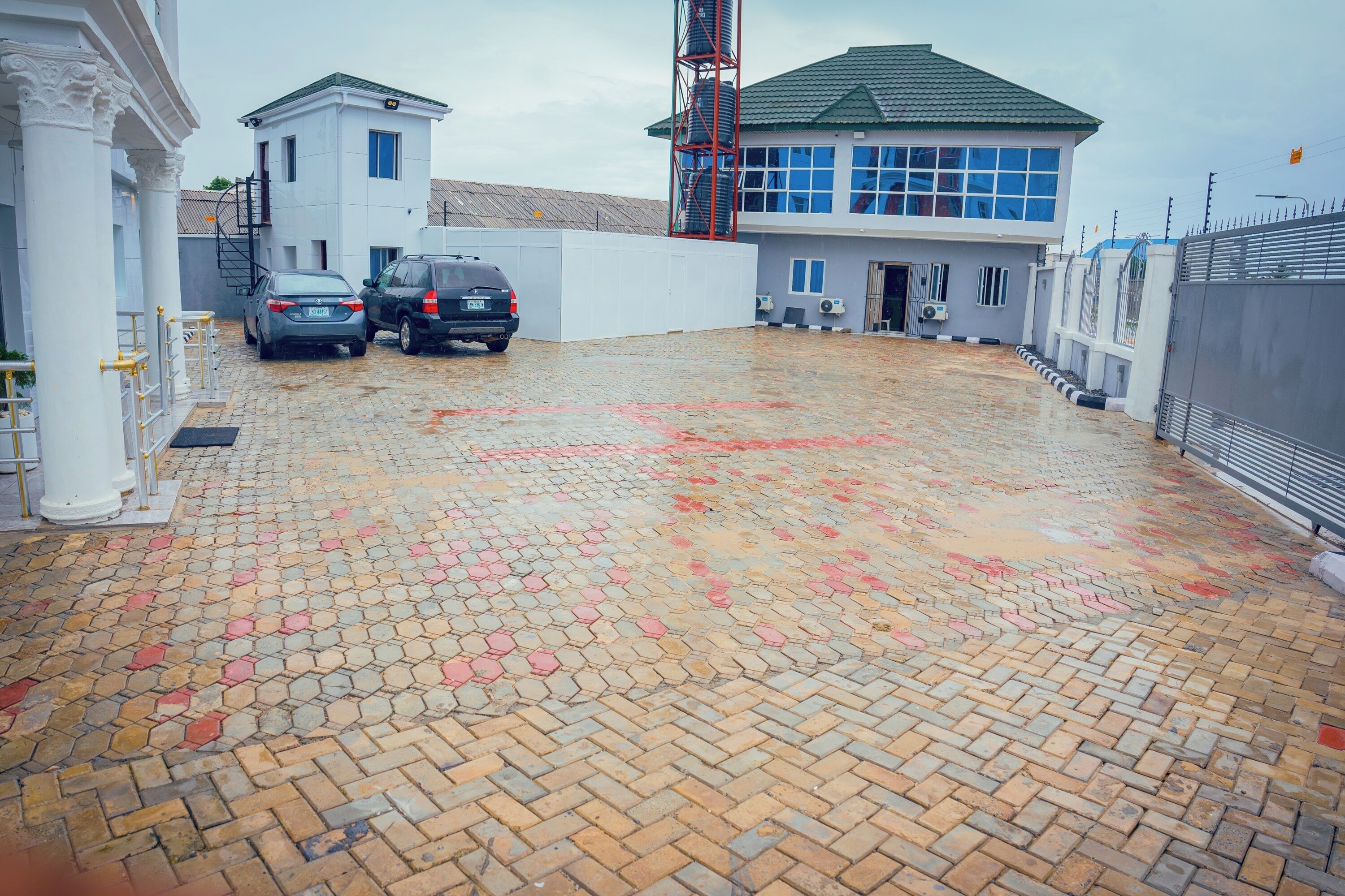 A paved courtyard with a brick floor, a white building with a blue window, and a metal fence.