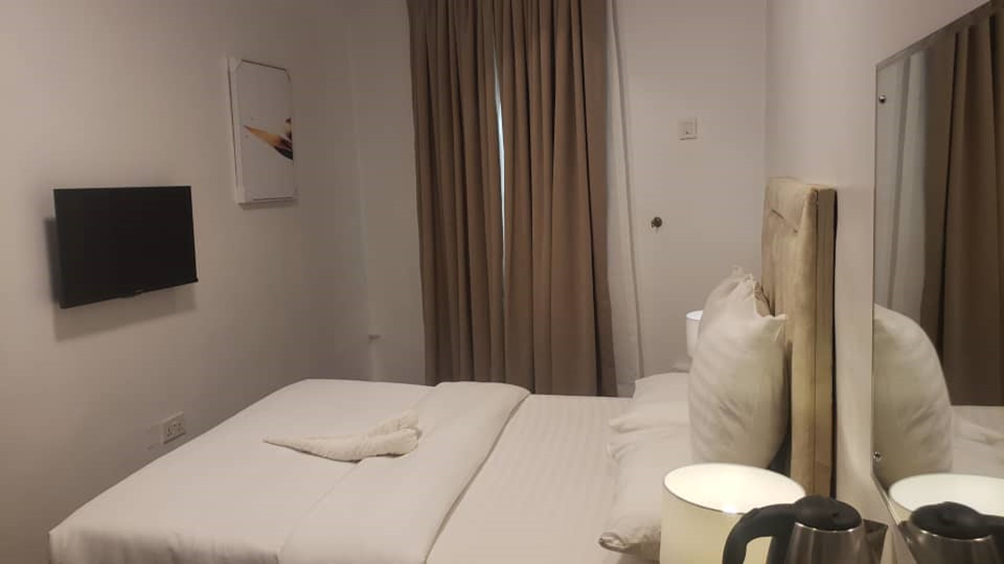 A hotel room with a bed, a flat-screen TV mounted on the wall, a window with curtains, a mirror, and a small table with a lamp.