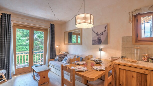 A cozy dining area with a wooden table and chairs, a bench, and a sofa. There is a large window with curtains, a ceiling light, and a wall-mounted picture of a deer. - Studio Montée du Calvaire - Welkeys (Megève)