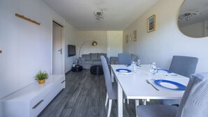 A modern, well-lit room with a dining table set for two, a sofa, and a small potted plant. - Appartement des Martrois - Welkeys (Villers-sur-Mer)