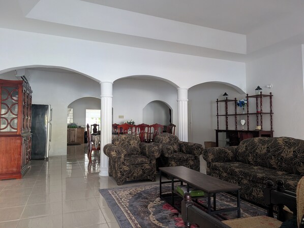 A spacious living room with a high ceiling, a dining area in the background, and a wooden cabinet with glass doors.
