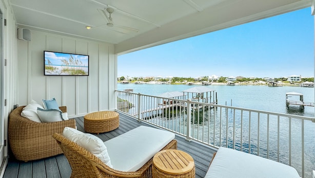 Ein Balkon mit Korbmöbeln, einem Flachbildfernseher und Blick auf Wasser und Gebäude.