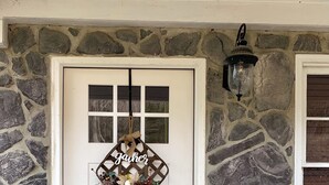 Una puerta blanca con un cartel de bienvenida en forma de diamante, una pared de piedra y un farolil encima.