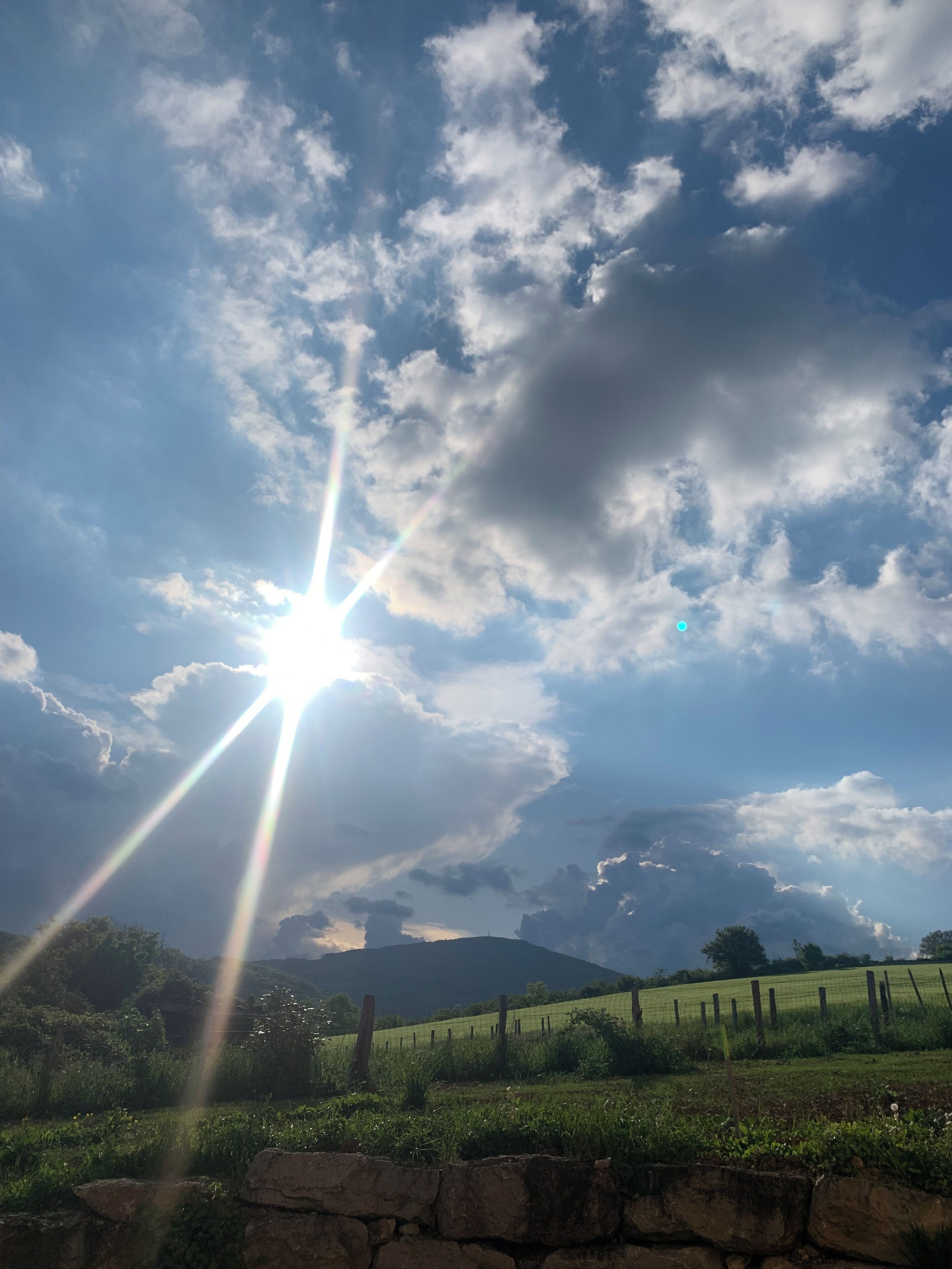 Paisagem rural com um muro de pedra em primeiro plano, um campo cercado e uma montanha ao longe, sob um céu parcialmente nublado com o sol brilhando.