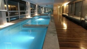 An indoor swimming pool with clear glass walls, wooden flooring, and a long, narrow layout.