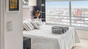 A bedroom with a large window, a bed with white bedding, and a view of the cityscape.