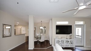 Un vaste espace de vie comprenant une table à manger, des meubles blancs, un téléviseur et un ventilateur de plafond.