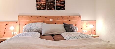 A bedroom with a wooden headboard, a bed with patterned bedding, two bedside lamps, and a wall-mounted picture of a sunset over a landscape.