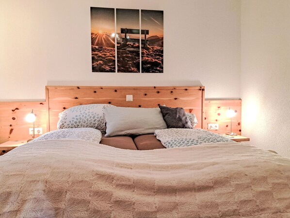 Une chambre à coucher avec une tête de lit en bois, un lit recouvert d’une literie à motifs, deux lampes de chevet et un tableau accroché au mur représentant un coucher de soleil sur un paysage.