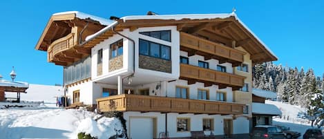 Ein mehrstöckiges Chalet mit Holzbalkonen, ein Sessellift im Hintergrund und eine verschneite Landschaft.