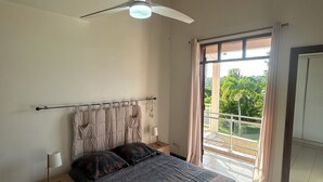 A bedroom with a bed, nightstand, lamp, ceiling fan, and a view of the outdoors through a large window. - Maison Sainte-anne (Sainte-Anne)