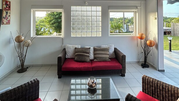A spacious living room with a high ceiling, a sofa with red cushions, two wicker chairs, and a glass coffee table. - Maison Sainte-anne (Sainte-Anne)