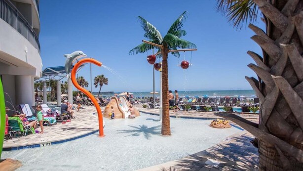 A pool area with a water slide, palm trees, and lounge chairs. - Oceans One 908 (Myrtle Beach)