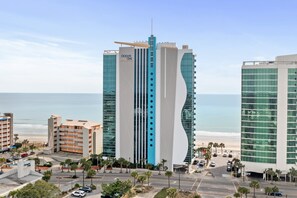 A modern hotel with a unique curved facade, a beach view, and a parking area with palm trees. - Oceans One 908 (Myrtle Beach)