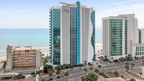 A modern hotel with a prominent vertical design, a beach view, and a parking area with palm trees. - Oceans One 908 (Myrtle Beach)