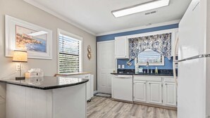 Une cuisine avec des meubles blancs, un plan de travail noir, une fenêtre avec des stores, une porte et un revêtement mural décoré d’un motif floral.