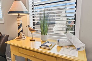 A hotel room with a desk, lamp, bird figurine, potted plant, notebook, towel, and parking permit.