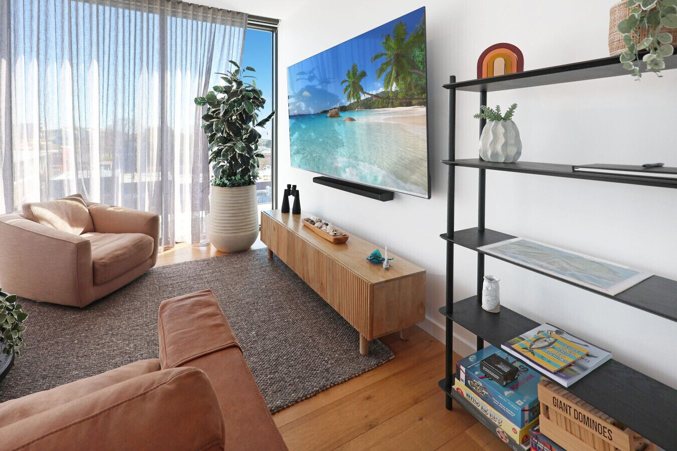 A modern living room with a beige sofa, a wooden coffee table, a large potted plant, a flat-screen TV mounted on the wall, and a bookshelf with various items.
