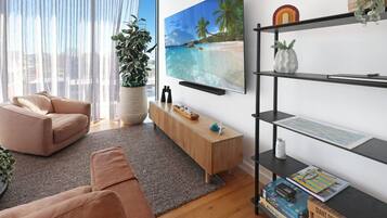 A modern living room with a beige sofa, a wooden coffee table, a large potted plant, a flat-screen TV mounted on the wall, and a bookshelf with various items.