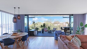 Un salon moderne doté d’une grande porte-fenêtre coulissante donnant sur un balcon aménagé avec des sièges et offrant une vue sur les arbres et les bâtiments.