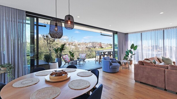Un salon moderne doté d’une grande porte vitrée donnant sur une terrasse, d’une table de salle à manger ronde avec des sets de table et d’un canapé agrémenté de coussins.