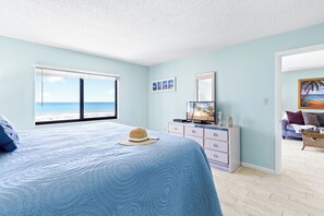 A bedroom with a large bed, a dresser, a television, and a view of the beach. - Direct Oceanfront Condo w/ Pool, Private Balcony & Ocean-View Master Bedroom (Cape Canaveral)