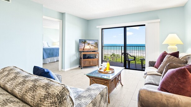 A hotel room with a balcony, a TV, a dining table with a bottle, and a sofa. - Direct Oceanfront Condo w/ Pool, Private Balcony & Ocean-View Master Bedroom (Cape Canaveral)