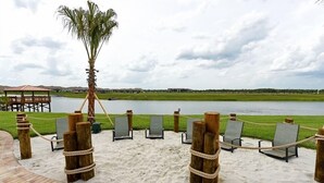 Zona arenosa de playa con sillas de descanso y una palmera, con vista a un cuerpo de agua y edificios a la distancia.