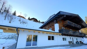 A two-story chalet with a wooden balcony, a stone foundation, and a snow-covered landscape.