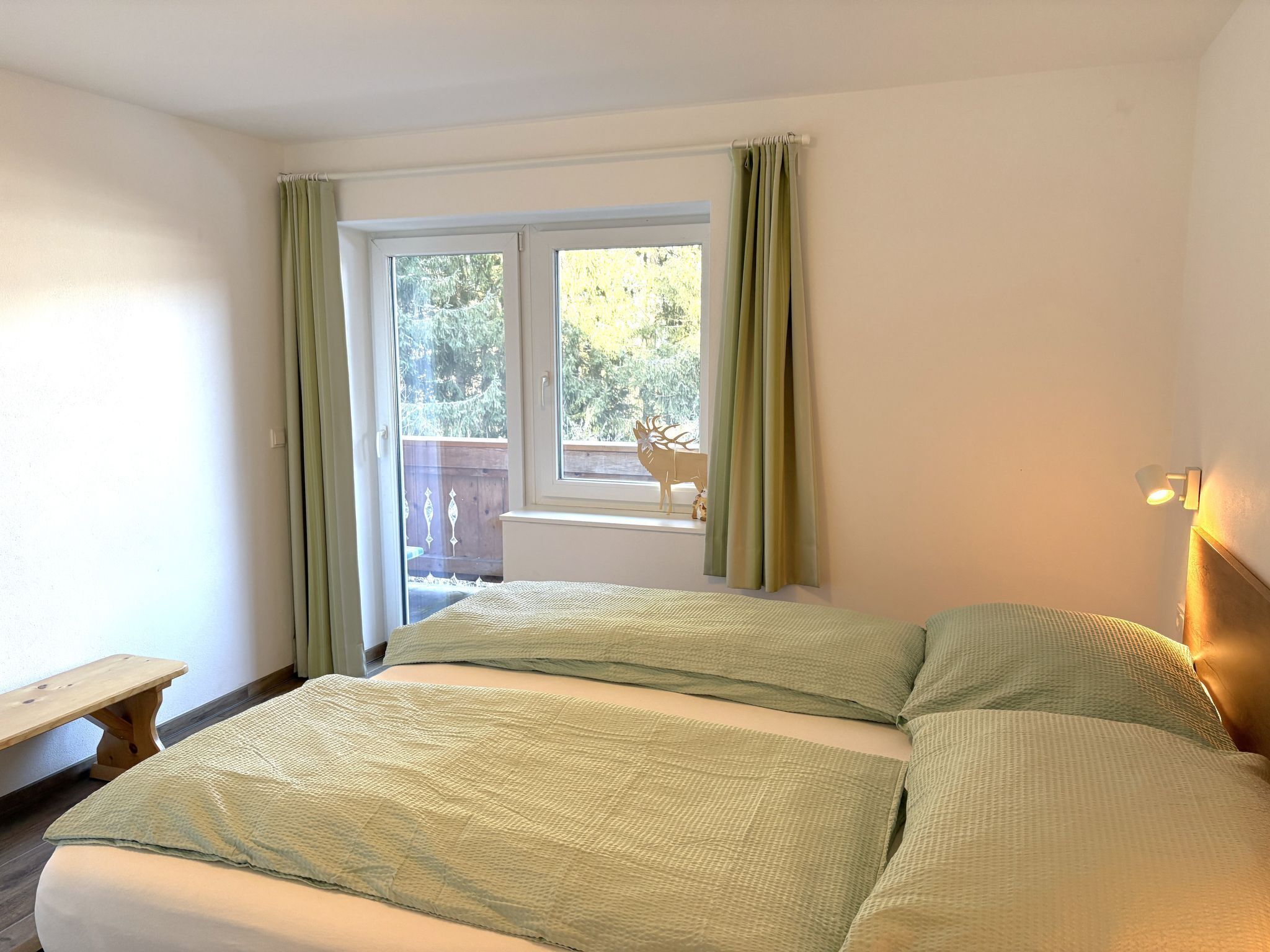 A bedroom with a bed, a bench, a window with curtains, and a view of the outdoors.