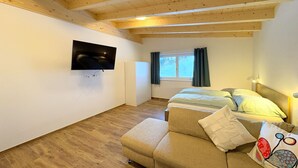 A room with a wooden ceiling, a bed with a headboard, a sofa, a TV mounted on the wall, and a window with curtains.
