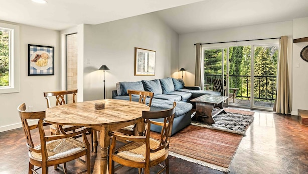 A modern living room with a wooden dining table, blue sofa, and a large sliding glass door leading to a balcony. - Bigwood Basecamp with Mountain Views & Great Location (Ketchum)