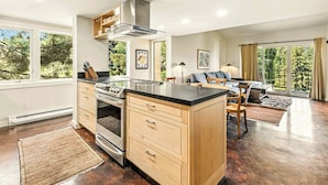 A modern kitchen with a large island, built-in oven, and stainless steel range hood. There is a dining area with a sofa and chairs, and a large window offering a view of trees. - Bigwood Basecamp with Mountain Views & Great Location (Ketchum)