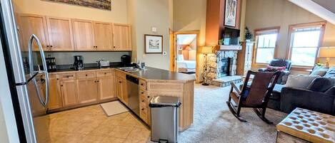 A spacious kitchen with wooden cabinets, a stainless steel refrigerator, and a small island. There is a trash can and a cozy seating area with a cushioned bench and a small table.