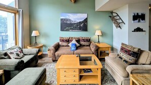 Un salon chaleureux avec une tête de cerf accrochée au mur, un canapé agrémenté de coussins à motifs, une table basse et un tableau représentant un paysage de montagne.