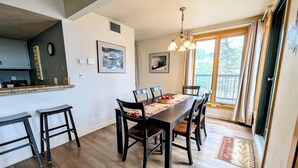 Un coin repas avec une table et des chaises en bois, un espace cuisine avec un comptoir et des tabourets, et une grande fenêtre avec des rideaux.