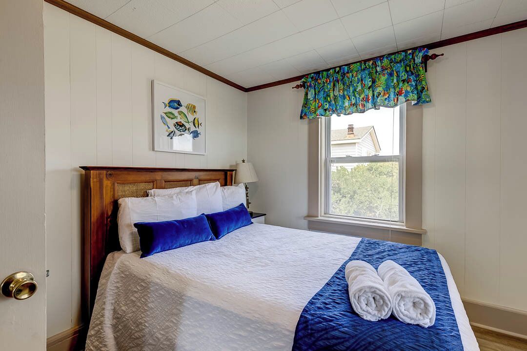 Een net opgemaakt bed met blauw-witte beddengoed, een houten hoofdbord en een raam met een kleurrijk gordijn.