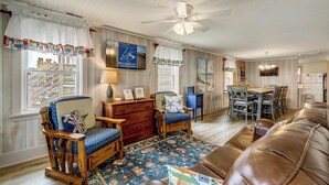A cozy living room with a wooden floor, a brown leather sofa, a wooden rocking chair, a blue patterned rug, and a dining table with chairs. - Breakers | Oceanfront (Nags Head)