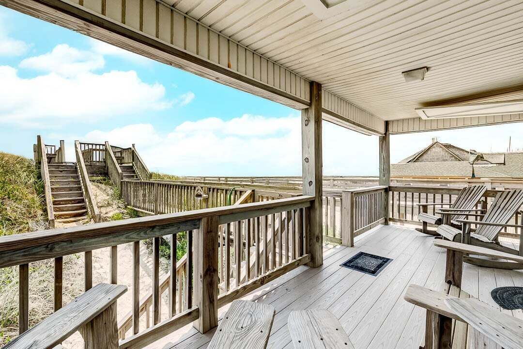 Een houten terras met een balustrade, een trap naar het strand en strandstoelen.