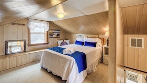 A bedroom with a wooden ceiling, a bed with blue and white bedding, a window with a view, and a small table with a lamp. - Breakers | Oceanfront (Nags Head)