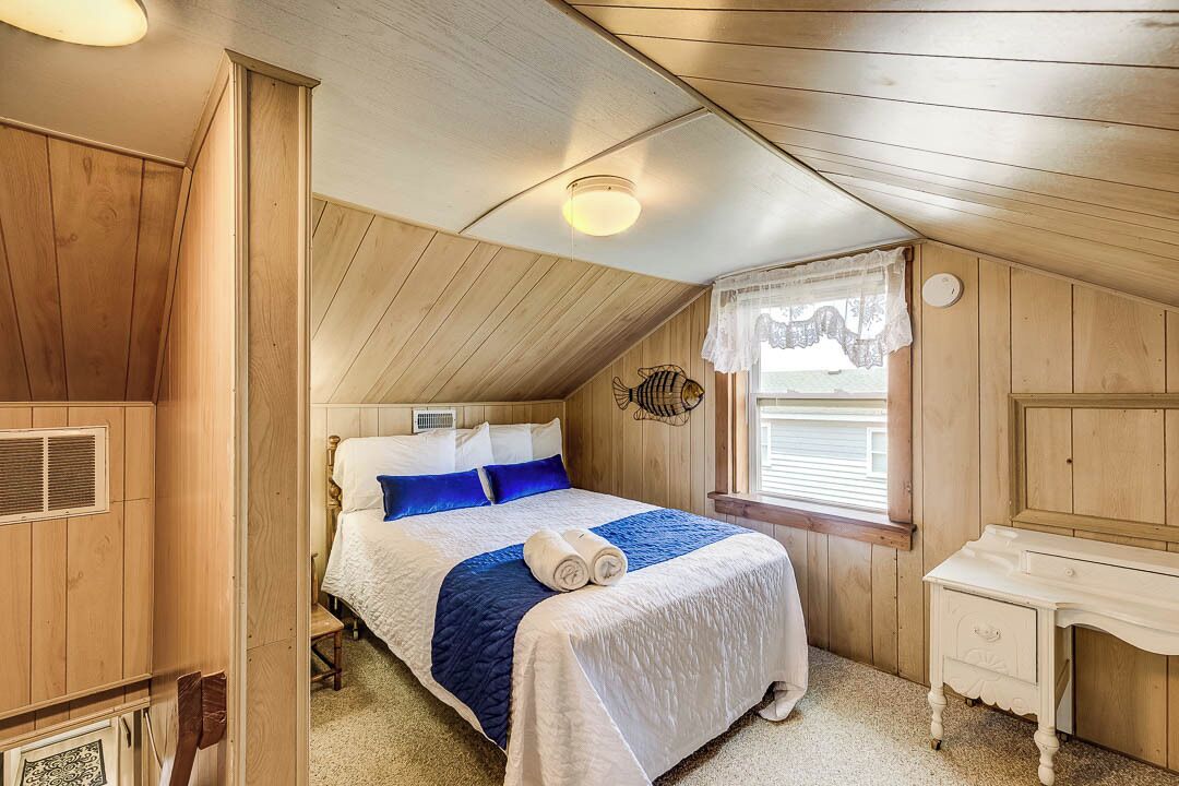 A cozy bedroom with a wooden ceiling, a bed with blue and white bedding, a bedside table, and a window with a lace curtain.