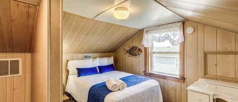 A cozy bedroom with a wooden ceiling, a bed with blue and white bedding, a bedside table, and a window with a lace curtain.