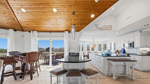 A spacious kitchen with white cabinetry, a central island with stools, and a dining area with wooden furniture. - Tide Together | 880 ft to the beach (Corolla)