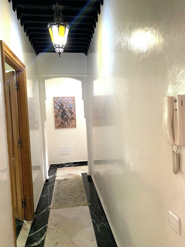 A hallway with a wooden door, a patterned rug, a marble floor, a wall-mounted phone, and a ceiling light fixture.